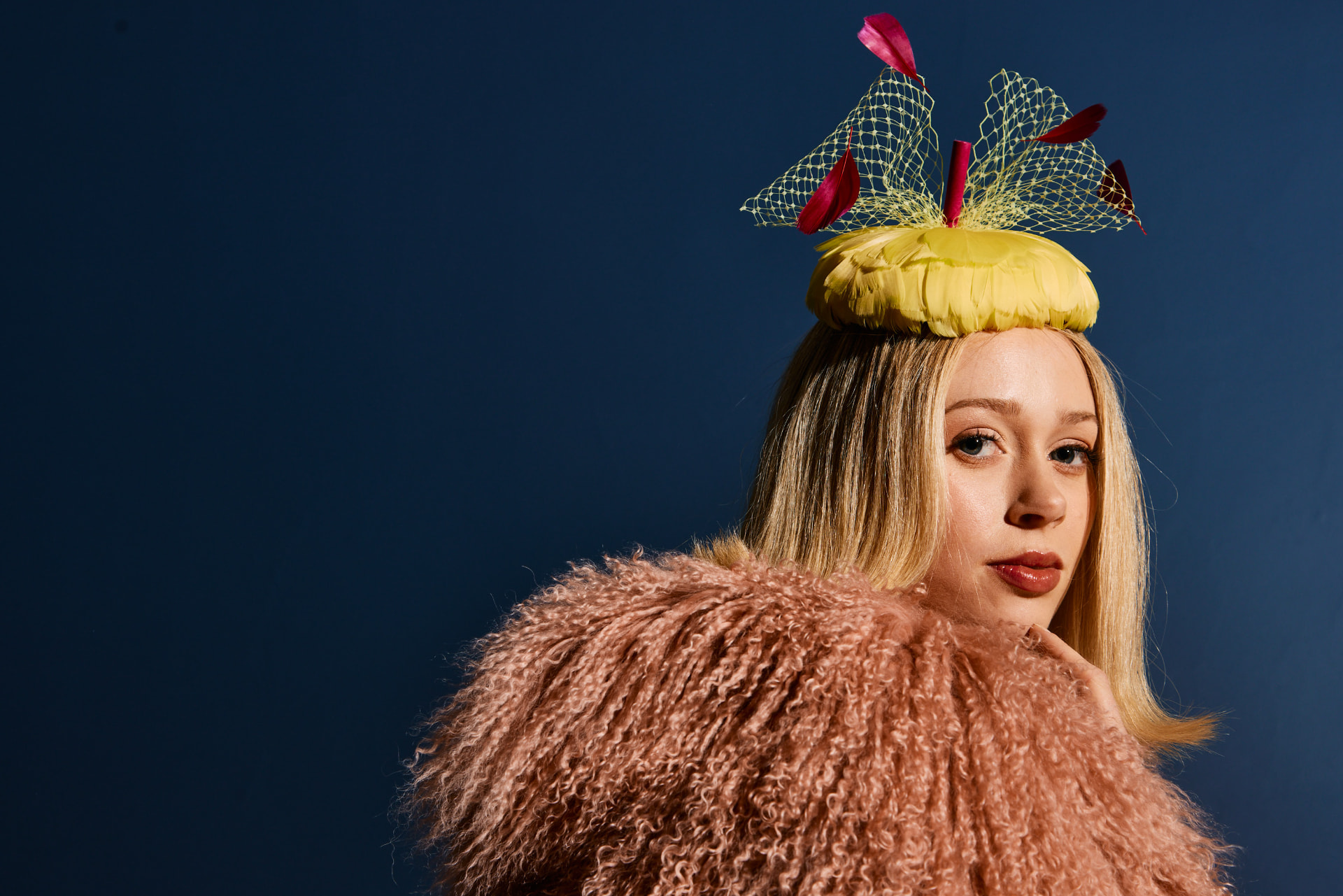 Model in a fluffy peach coat wearing a yellow feathered percher with scarlet quills and veiling, against a deep navy backdrop